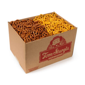 Open cardboard box containing small brown pretzels on the left and small yellow (cheese) pretzels on the right. The side of the box has red printed text and a logo.
